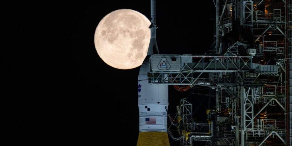 NASA Artemis SLS rocket on the launch pad at night with a full Moon rising behind it, showing the NASA logo and American flag on the spacecraft
