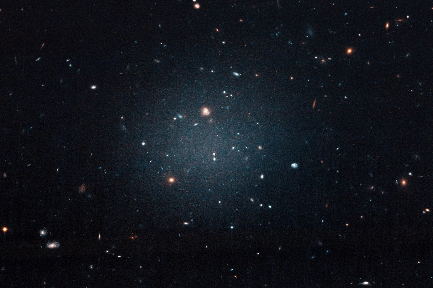 Hubble Space Telescope image of the ultra-diffuse galaxy NGC 1052-DF2, a faint ghostly glow with distant galaxies visible through it — the first galaxy found lacking dark.