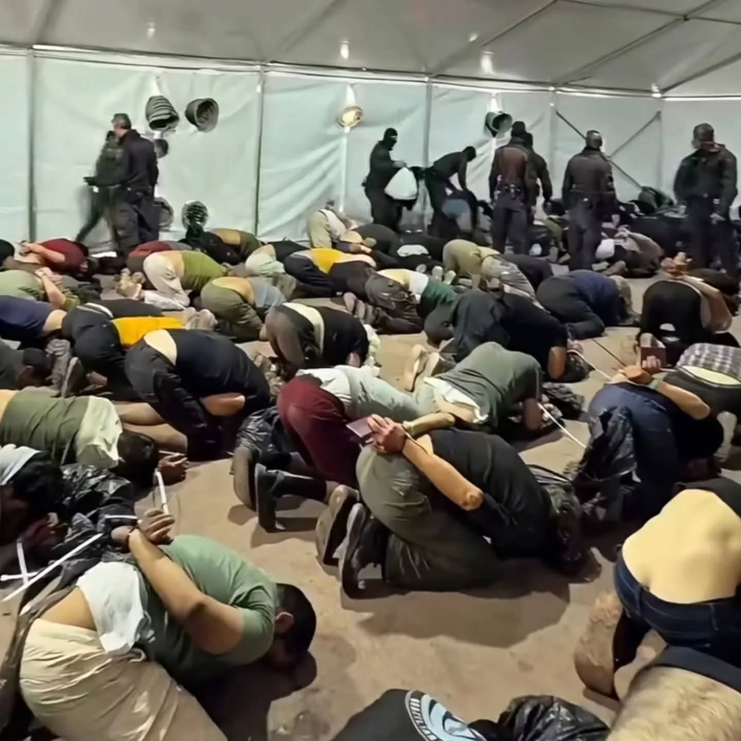 Handcuffed Flotilla activists kneeling on the ground inside a tent in Ashdod, Israel, surrounded by armed personnel after being detained in international waters.