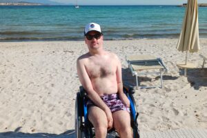 Gerd Dani in a wheelchair on a wooden accessibility walkway at Fertilia beach, Sardinia, with the turquoise Mediterranean sea, a sailboat, and distant Alghero coastline behind him.