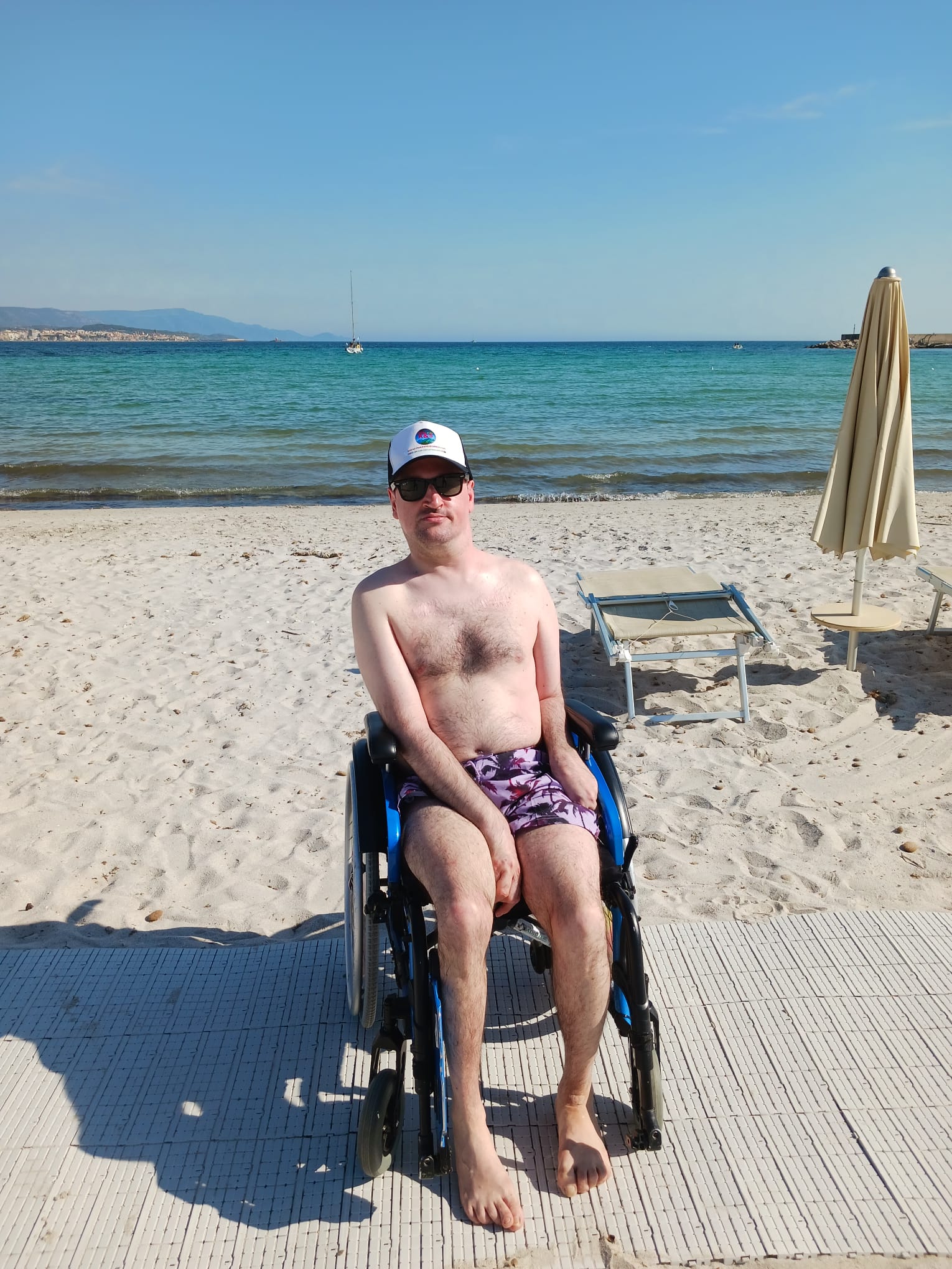Gerd Dani in a wheelchair on a wooden accessibility walkway at Fertilia beach, Sardinia, with the turquoise Mediterranean sea, a sailboat, and distant Alghero coastline behind him.