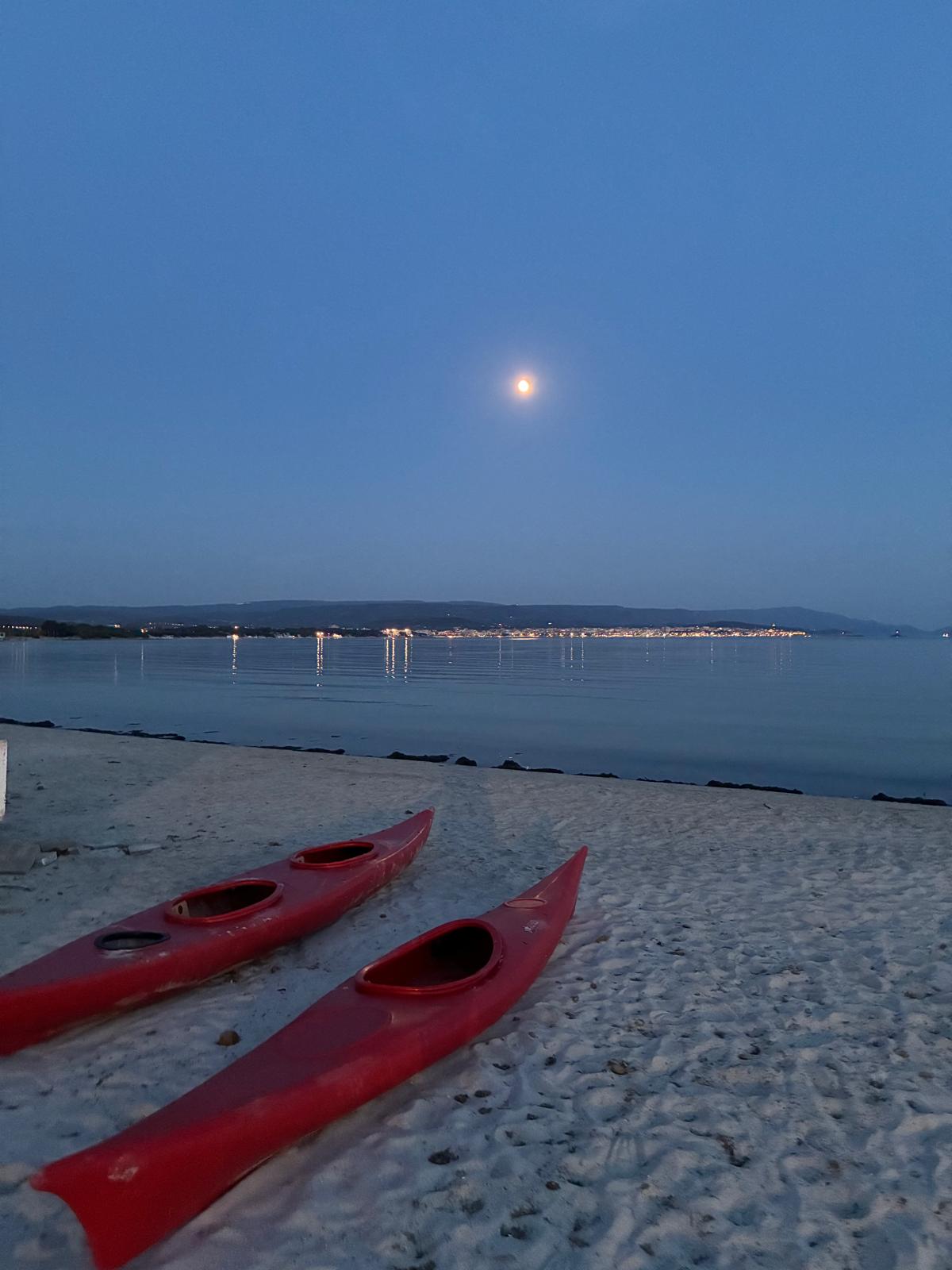 Full moon rising over Fertilia bay at dusk in Sardinia, with two red kayaks resting on white sand and the lights of Alghero shimmering across calm sea.