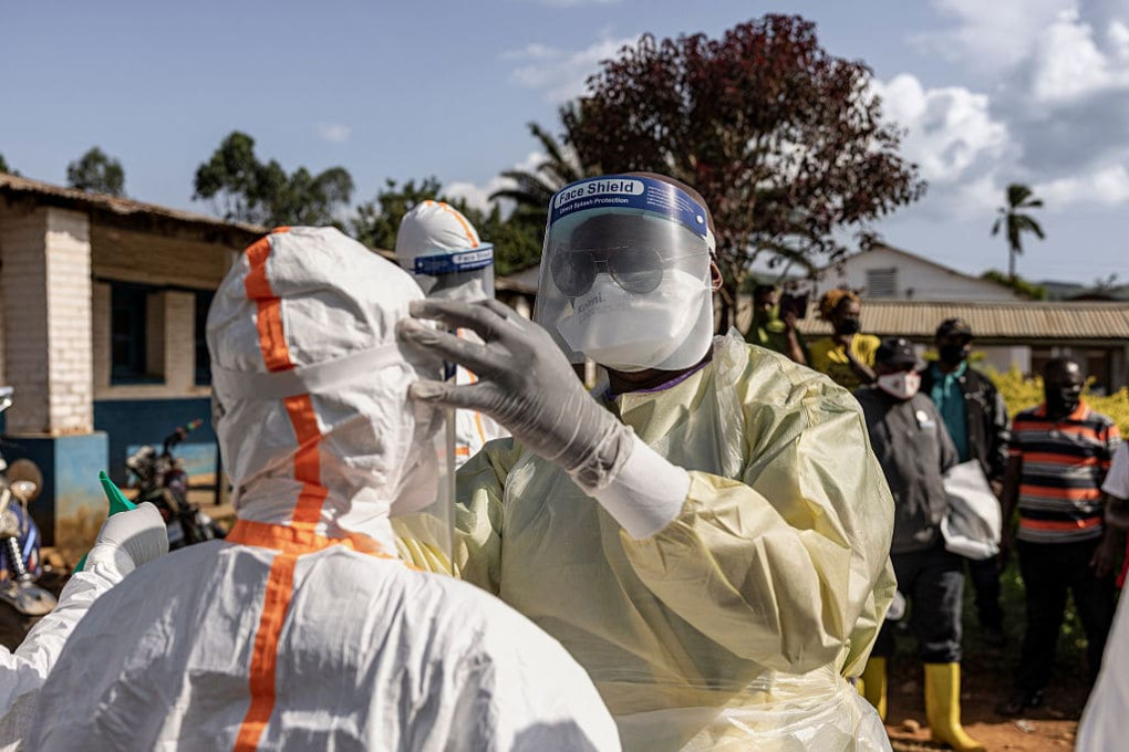 Two health workers in white PPE suits and face shields verify protective equipment outside an Ebola treatment hospital in Mongbwalu, Ituri, DRC, May 2026.