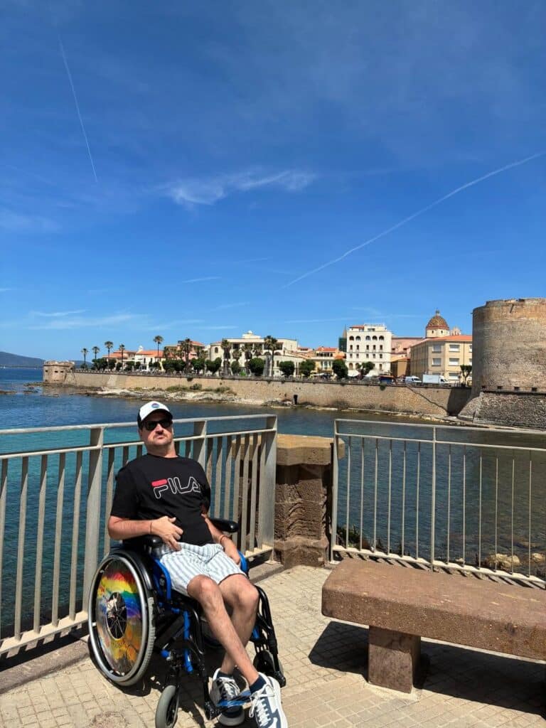 Me in wheelchair on Alghero seafront promenade with Sardinia's medieval bastions, turquoise sea, and historic Old Town in the background.