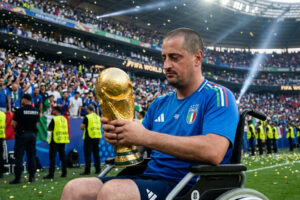 Man in Italy Azzurri jersey holds golden FIFA World Cup trophy from his wheelchair as fans wave Italian flags in the stadium behind him.