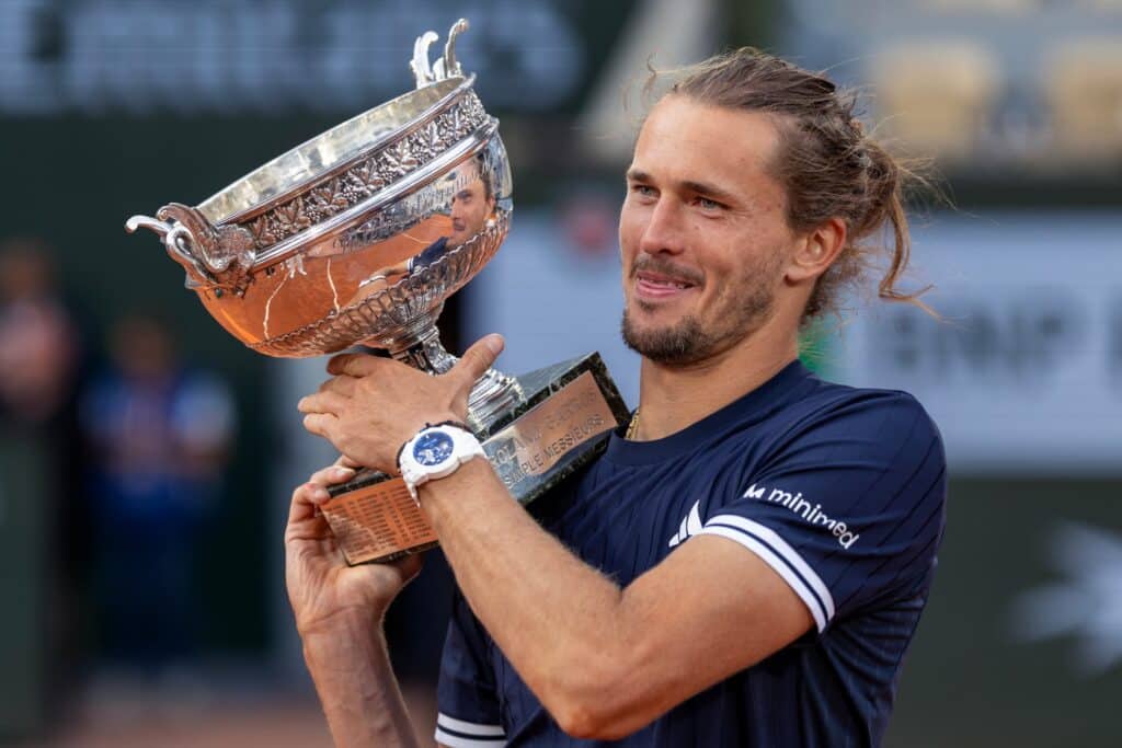 Alexander Zverev smiling while holding the Roland Garros 2026 Coupe des Mousquetaires trophy, wearing a navy Adidas shirt with MiniMed diabetes-care sponsor logo.
